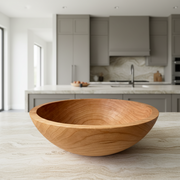 A round wooden bowl with a smooth, polished finish and visible wood grain, resting on a light stone surface against a neutral gray wall.