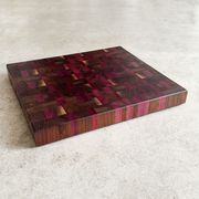 A rectangular wooden cutting board with a complex geometric inlay pattern of dark brown, reddish-brown, and pink wood tones, shown on a light gray concrete surface.