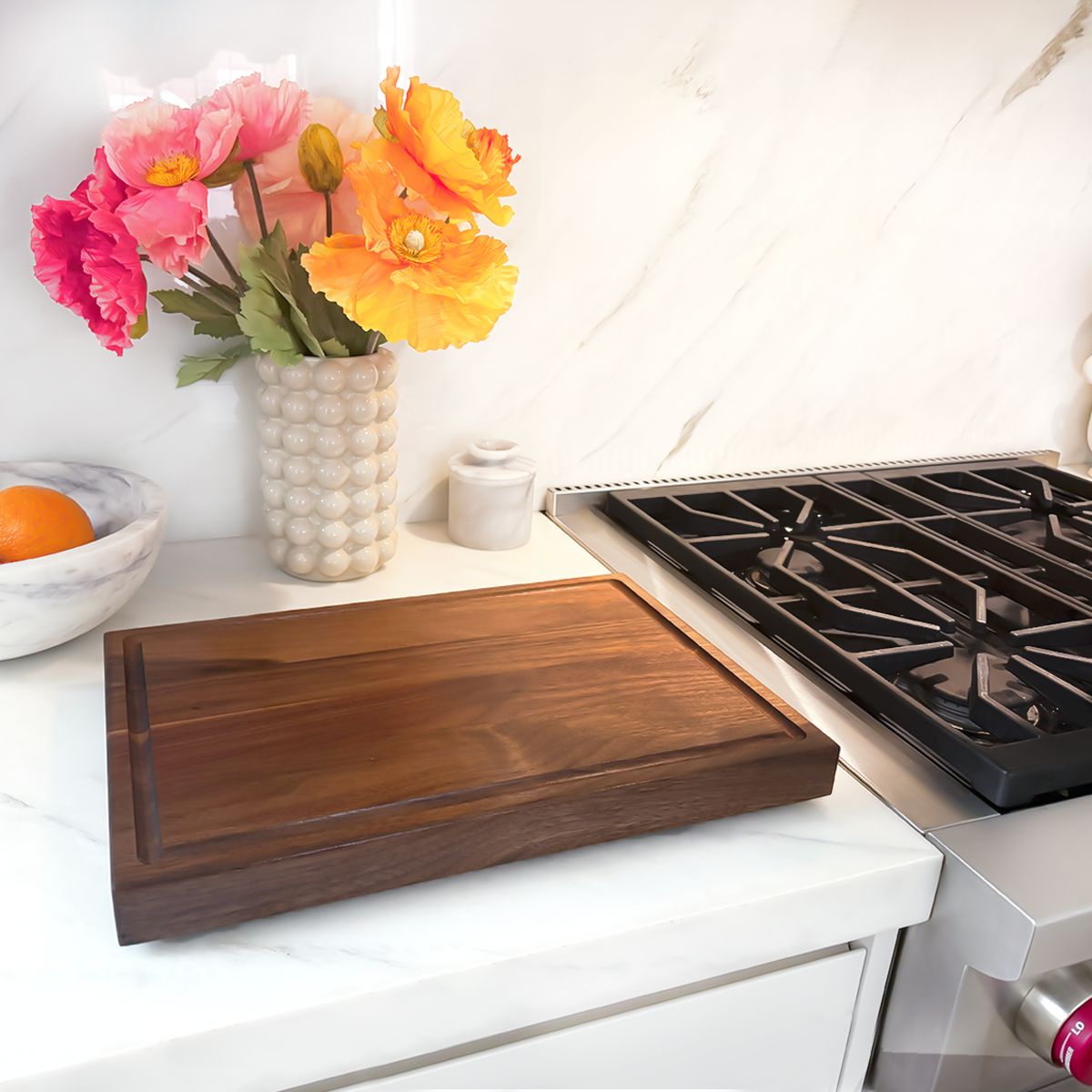 Wooden cutting board on a kitchen counter with flowers and oranges in the background